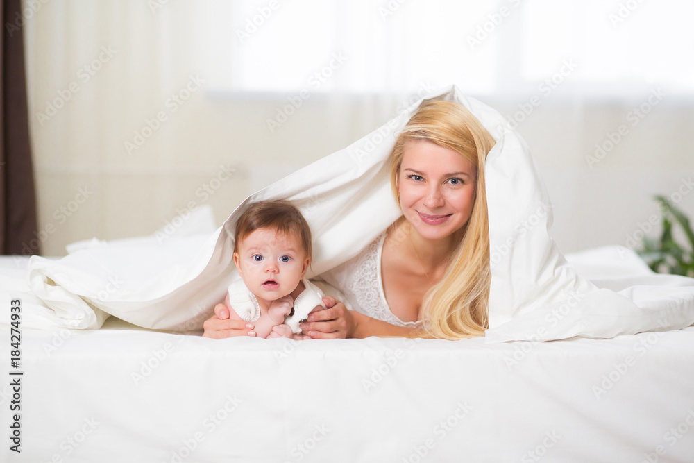 Portrait of young happy smiling mother with infant baby girl liying on the bed covered with a white blanket. Happy family concept Portrait of young happy smiling mother with infant baby girl liying on the bed covered with a white blanket. Happy family concept