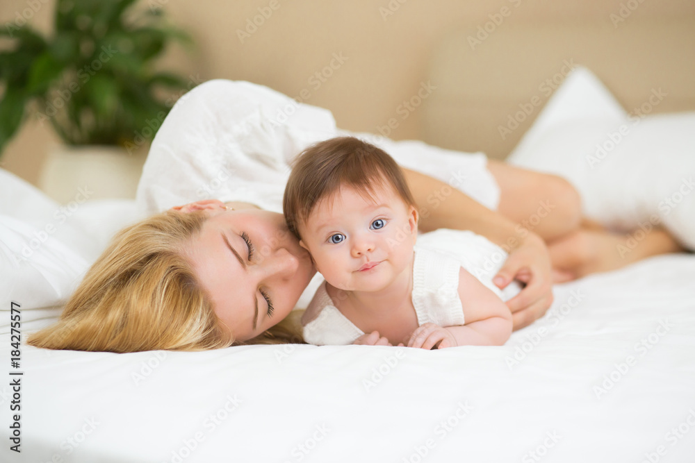 Portrait of young mother with blond hair with her sweet 3 month old baby girl in white wear having fun in the bedroom at morning, loving happy family concept Portrait of young mother with blond hair with her sweet 3 month old baby girl in white wear having fun in the bedroom at morning, loving happy family concept