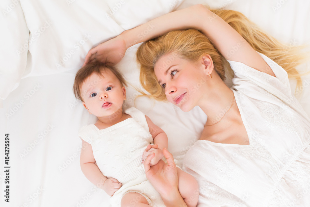 Happy loving family concept. Beautiful mother playing with her baby girl in the bedroom. They smiling and hugging together on white bed linens, top view Happy loving family concept. Beautiful mother playing with her baby girl in the bedroom. They smiling and hugging together on white bed linens, top view