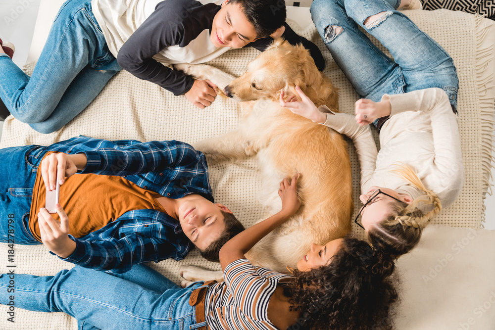 overhead view of multiethnic teenagers lying on bed and taking selfie with dog overhead view of multiethnic teenagers lying on bed and taking selfie with dog