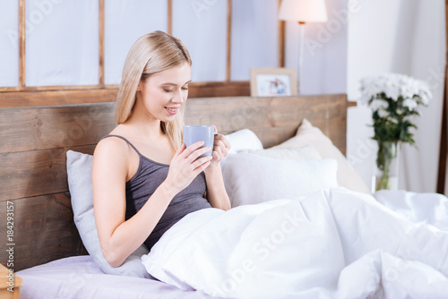 Delicious drink. Charming young woman sitting in her bed under the blanket and drinking a big mug of coffee