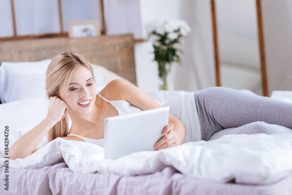 New discoveries. Charming fair-haired young woman lying on the bed and reading news from her tablet while resting her cheek on the hand New discoveries. Charming fair-haired young woman lying on the bed and reading news from her tablet while resting her cheek on the hand