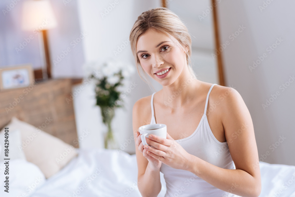 Sweet drink. Cheerful pretty woman standing in the bedroom and posing for the camera while holding a cup of tea Sweet drink. Cheerful pretty woman standing in the bedroom and posing for the camera while holding a cup of tea