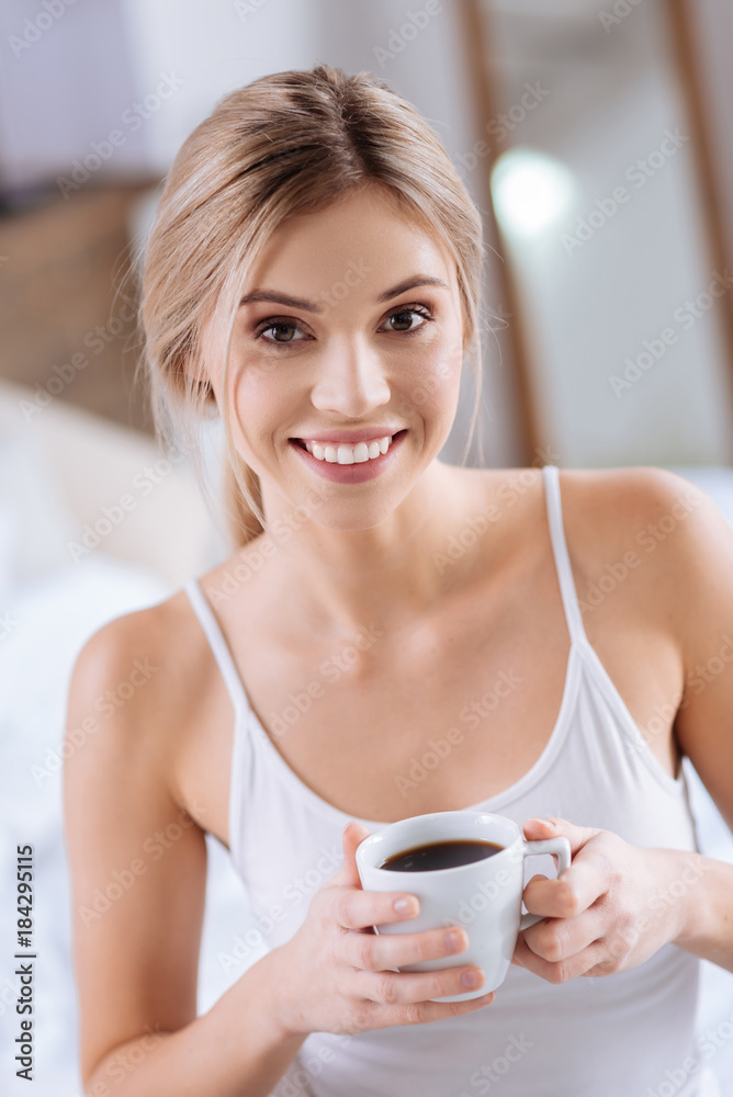 Enjoy this drink. The portrait of a charming slender young woman holding a cup of coffee and smiling at the camera Enjoy this drink. The portrait of a charming slender young woman holding a cup of coffee and smiling at the camera