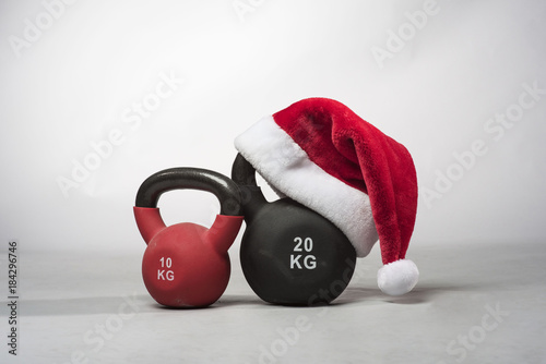 A large black Kettlebell wearing a red  Santa hat next to a smaller red kettle bell on a plain light grey background 