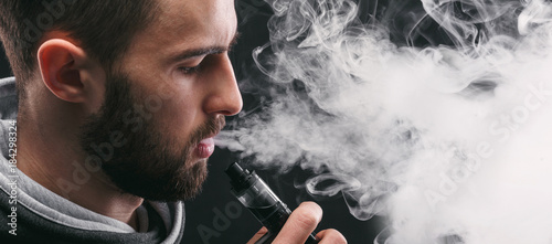 Young man vaping e-cigarette with smoke on black photo