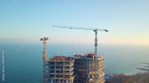 Aerial city view. Construction of a high-rise skyscraper on the ocean by two cranes. Even slow fly. The camera focusses the view on the high-rise crane and turns down.