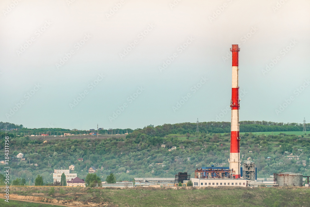 Factory chimney under the green hill. Dusty industrial landscape.