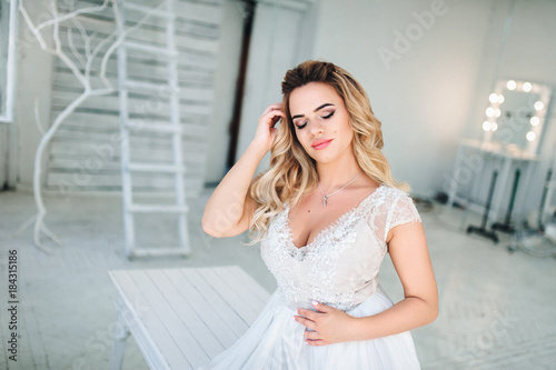 Attractive blonde bride is standing by the window in the white interior with her eyes closed. Sexy bride. Interior in vintage style.