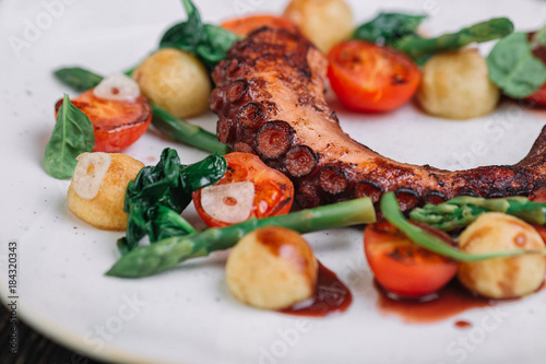 Fried octopus tentacles with vegetable on white plate on dark wood background