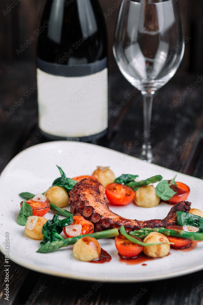 Fried octopus tentacles with wineglass and bottle of wine on white plate on dark wood background
