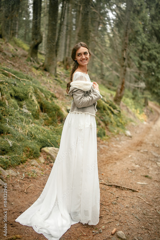 Bride stands on forest road, smiles and looks back.