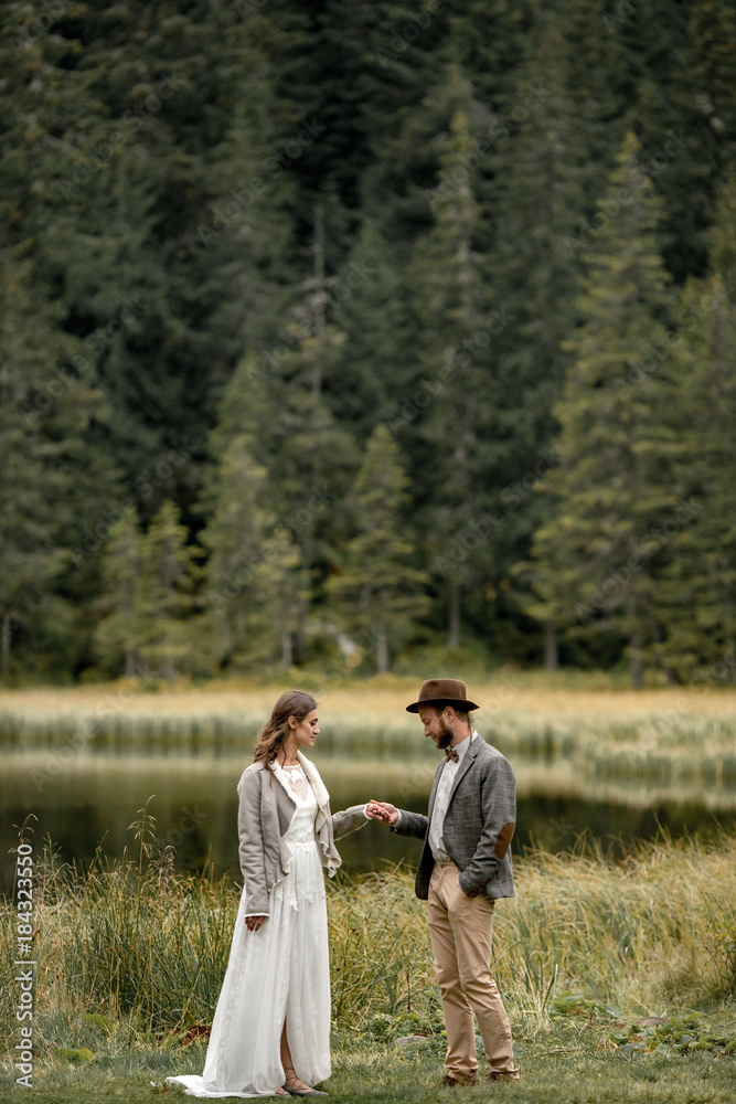 Bride and groom are standing and holding hands near forest lake.