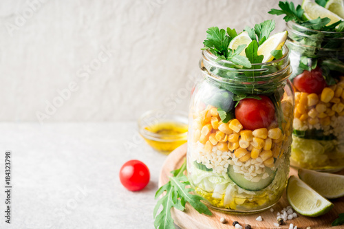 Healthy homemade salads with bulgur, vegetables and lime in mason jars on grey stone background.