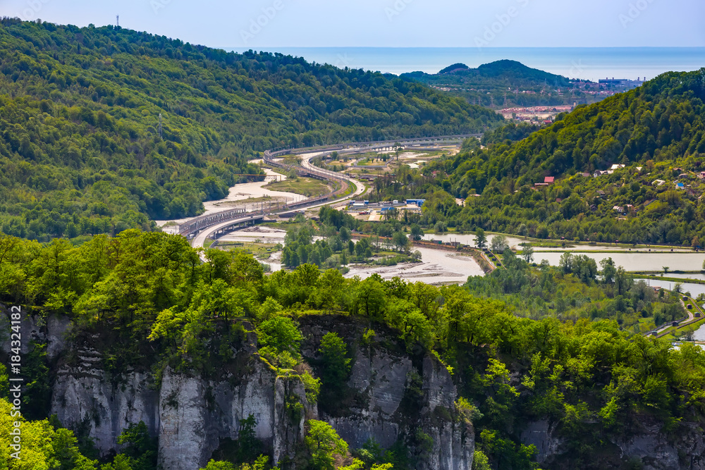 High-speed highway next to a rocky mountain with a green spring forest around a trout fishermen's farm in the background of the Black Sea on the horizon. The road Adler Krasnaya Polyana, Sochi Russia.