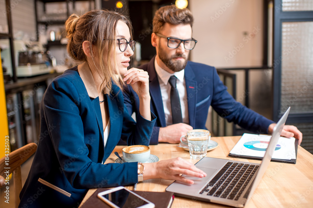 Caucasian business couple dressed strictly in the suits working together with laptop and paper charts sitting at the cafe during the coffee break Caucasian business couple dressed strictly in the suits working together with laptop and paper charts sitting at the cafe during the coffee break