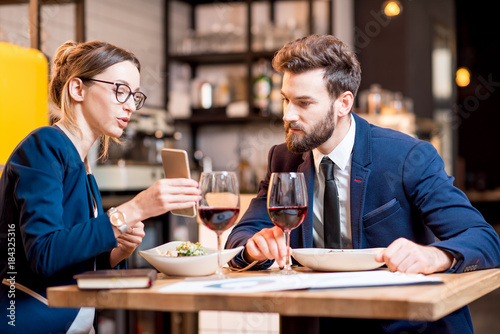 Caucasian business couple dressed strictly in the suits working together with smart phone sitting at the restaurant during the dinner photo