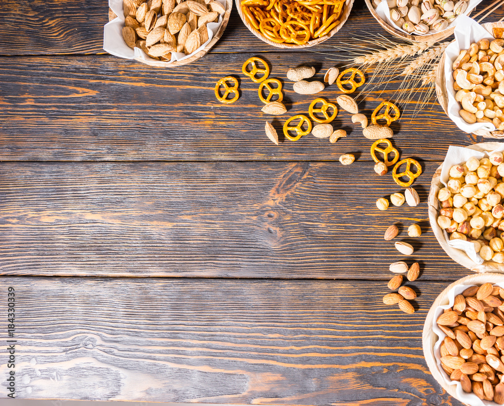 Top view of various beer snacks in plates like pistachios, small pretzels and different peanuts on dark wooden desk