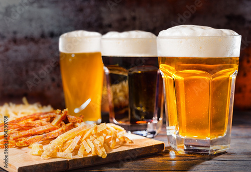 Three glasses with freshly poured light  unfiltered and dark beer stand near wooden cutting board with dried fish on dark desk