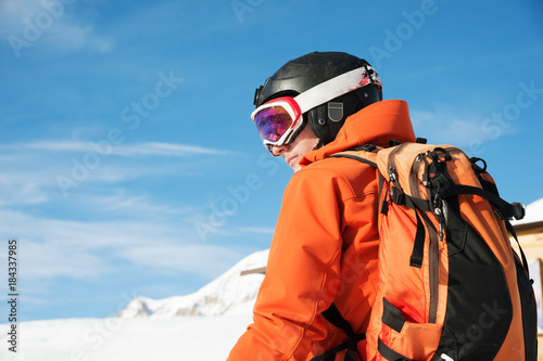 Portrait of a skier in an orange overall with a backpack on his back in a helmet stands against the background of a beautiful Caucasian mountain landscape