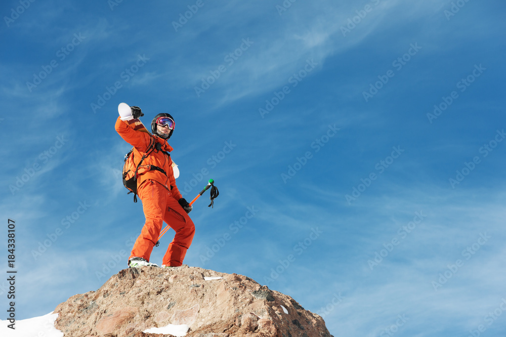 Portrait of a Skier in an orange overall with a backpack on his back and skis on his shoulders in a helmet stands on a rock against the blue sky.