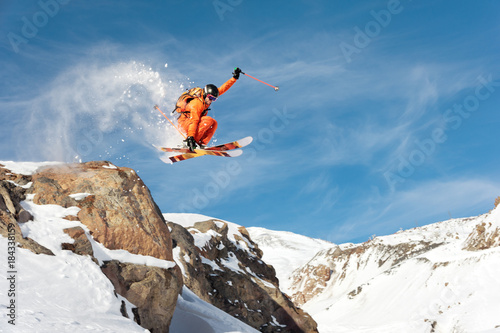 A professional skier makes a jump-drop from a high cliff against a blue sky leaving a trail of snow powder in the mountains