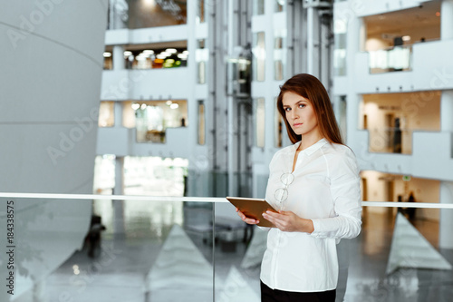 The business woman in a fair hair in white office shirt works on the tablet and checks information on the Internet. The girl costs in office building against the background of business center.