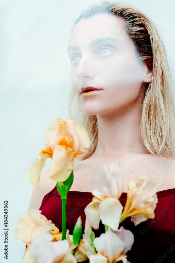 portrait of the young blonde attractive woman with the fashion alien gold make up standing with the iris flowers behind the glass