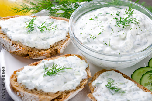 Homemade greek tzatziki sauce in a glass bowl with ingredients and sliced bread on a dark black stone background. Cucumber, lemon, dill, garlic. Close-up, horizontal, selective focus on a bowl photo
