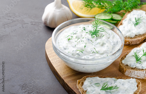 Homemade greek tzatziki sauce in a glass bowl with ingredients and sliced bread on a dark black stone background. Cucumber, lemon, dill, garlic. Close-up, horizontal, selective focus on a bowl
