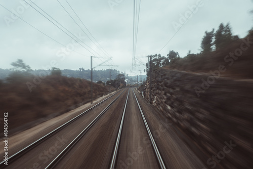 Photo shooting from the last coach of a high-speed train: dry autumn landscape with dull sky, two railway tracks stretching into distance, stone wall, plants and trees, poles and wires