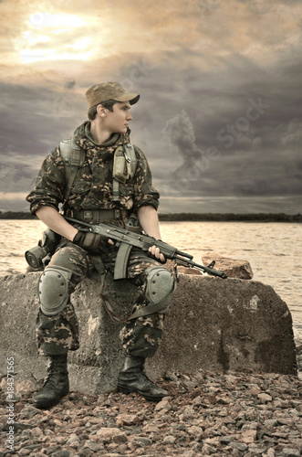 young soldier with a gun sitting on a rock near water