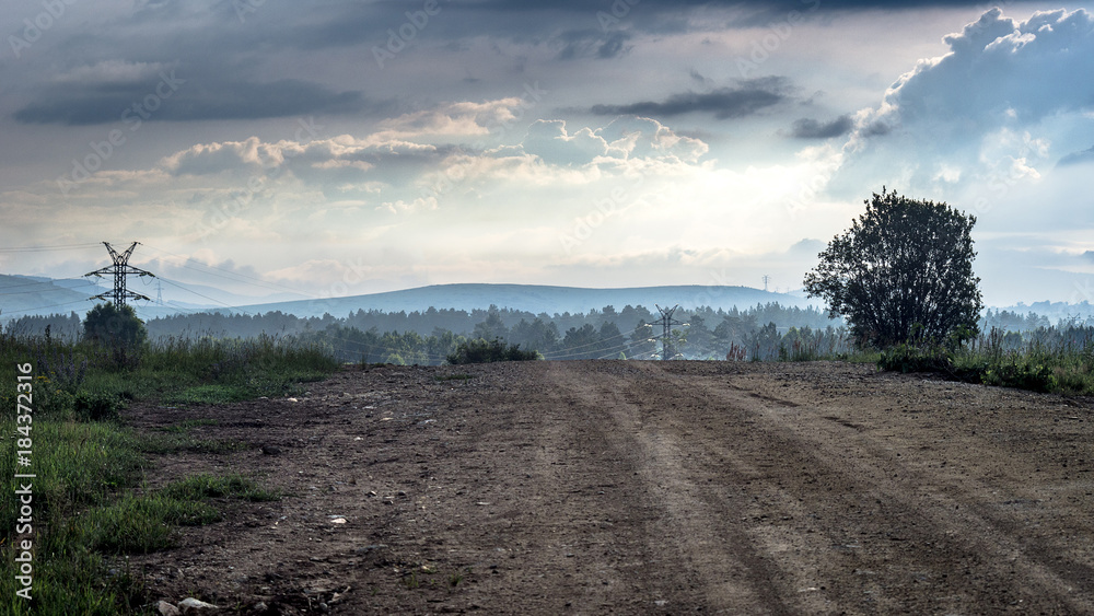 Fototapeta premium Rural mountain country road at misty summer morning with dramatic clouds and power lines