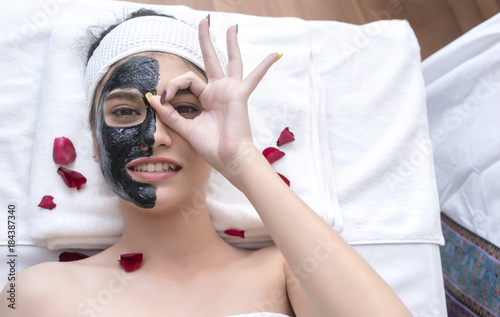 Beautiful smiling young Asian women having revitalizing mud mask and giving Ok sign , lying on massage table in spa salon