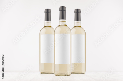 Three trasparent wine bottles with blank white labels on white wooden board, mock up. Template for advertising, design, branding identity.