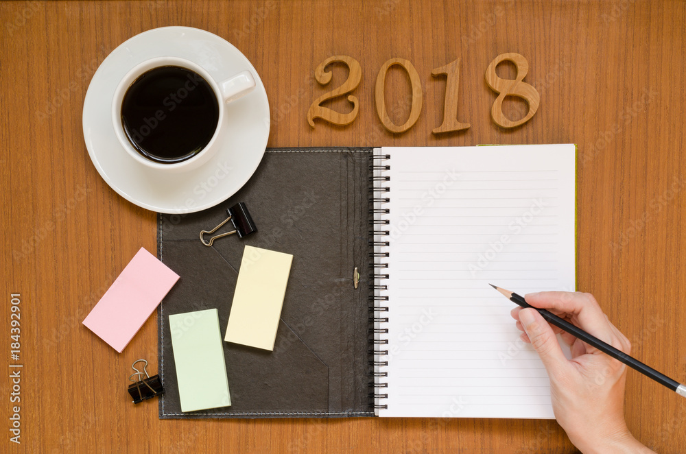 Recycled notebook with woman hand writing in the empty paper note surrounding with black coffee in white cup, 2018 letters, yellow, pink, green sticky pad, pencil & paper clip on wood background space Recycled notebook with woman hand writing in the empty paper note surrounding with black coffee in white cup, 2018 letters, yellow, pink, green sticky pad, pencil & paper clip on wood background space
