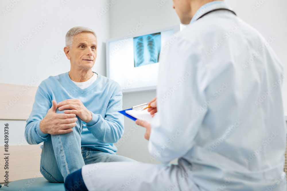 Obraz premium Pain in the knee. Good looking nice aged man holding his knee and looking at his doctor while explaining where he feels pain