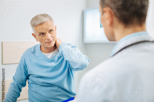 Unpleasant feelings. Nice good looking aged man looking at his doctor and holding his neck while feeling pain
