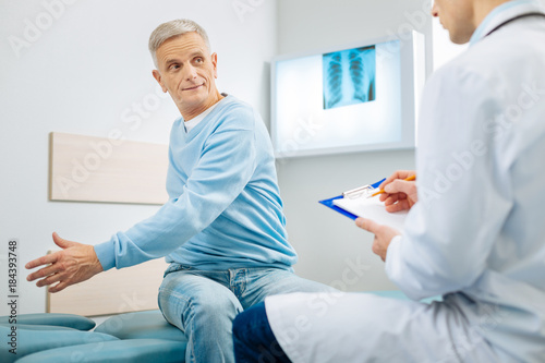 Caring about health. Delighted nice senior man sitting on the bed and turning his body while visiting a doctor