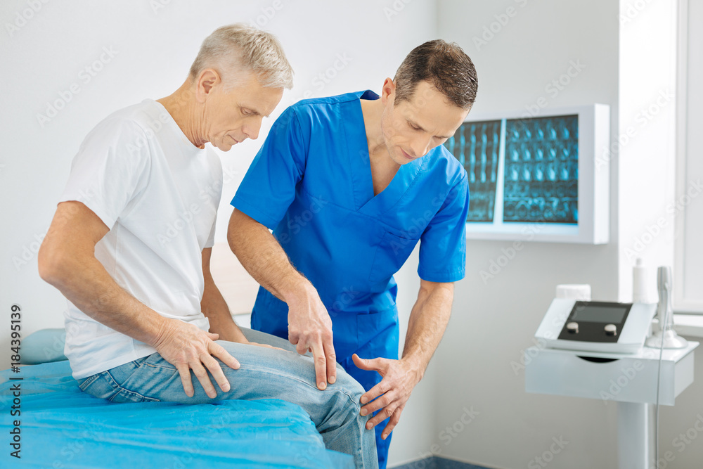 Fototapeta premium Absolutely concentrated. Serious pleasant male therapist standing near his patient and checking his leg while focusing on his work