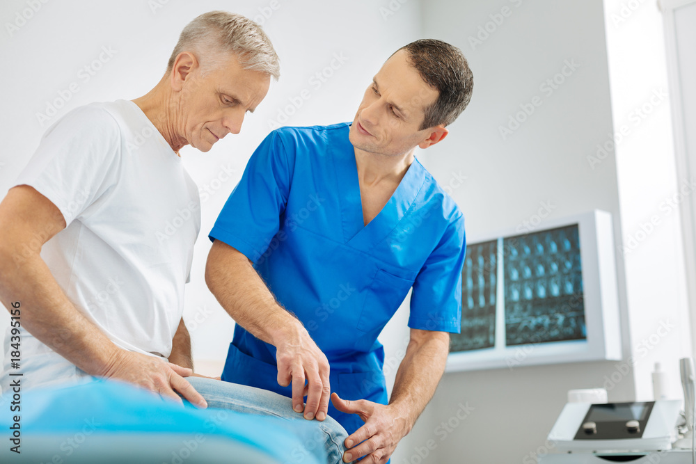 Diagnostic procedure. Smart pleasant skillful doctor standing near his patient and pressing his finger to his knee while doing medical diagnostics Diagnostic procedure. Smart pleasant skillful doctor standing near his patient and pressing his finger to his knee while doing medical diagnostics