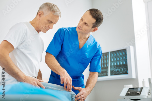 Diagnostic procedure. Smart pleasant skillful doctor standing near his patient and pressing his finger to his knee while doing medical diagnostics