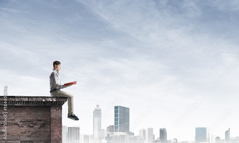 Man on roof edge reading book and cityscape at background