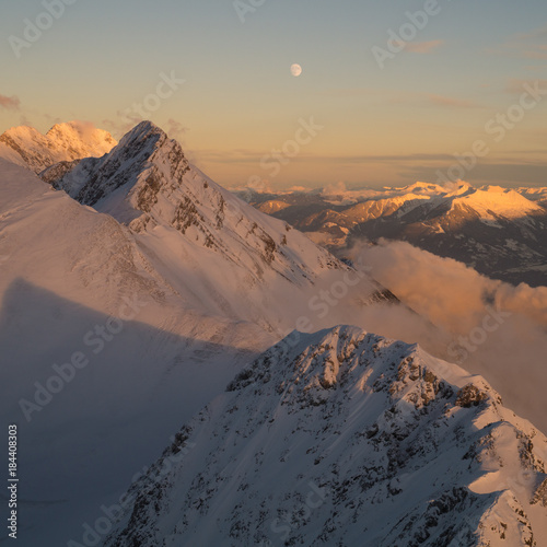 Clouds making their way up a mountain ridge during a winter sunset with the moon in the background.