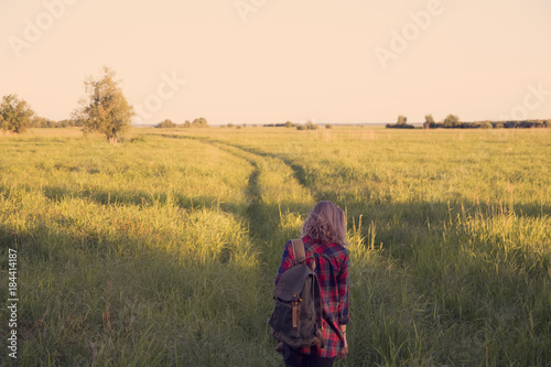 girl in plaid shirt with a backpack on shoulders walking down country road in the middle of a field with tall grass © UlianaG