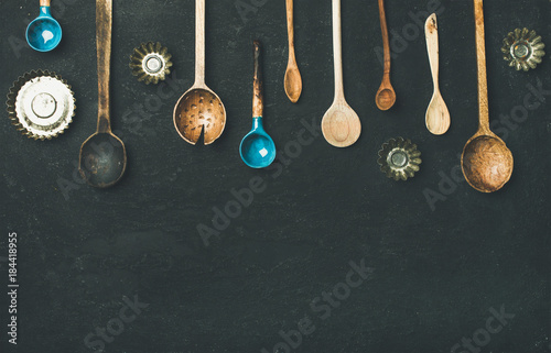 Flat-lay of various old vintage kitchen spoons and baking tin molds over black stone background, top view, copy space. Rustic cooking concept photo