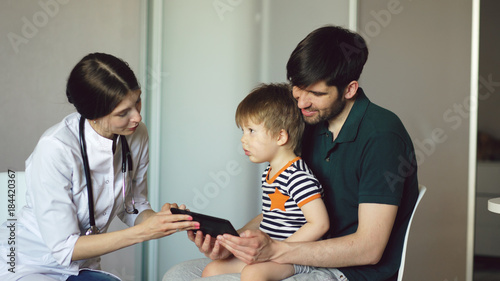 Young woman doctor talking with father of little boy using tablet computer in medical office