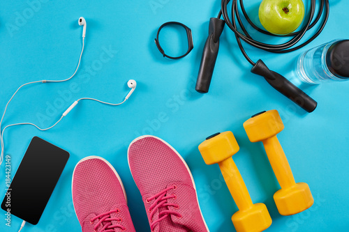 Flat lay shot of sneakers, jumping rope, dumbbells and smartphone on blue background. © nazarovsergey