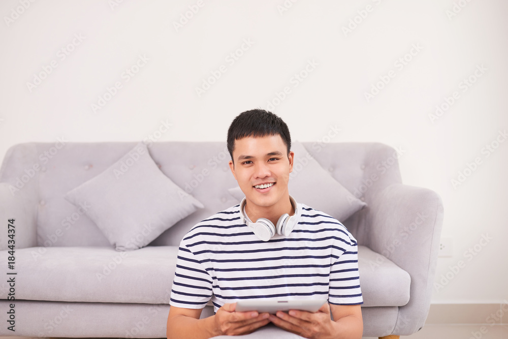 Handsome guy is using a digital tablet, looking at camera and smiling while sitting on the floor at home Handsome guy is using a digital tablet, looking at camera and smiling while sitting on the floor at home
