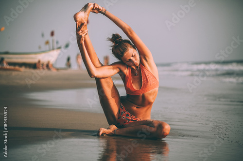 young flexible woman in swimsuit practicing yoga on beach at goa